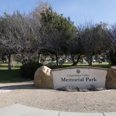 A sign reading Carpinteria Valley Memorial Park is flanked by boulders, with trees and a grassy area in the background.