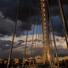 A bridge with sleek, modern cables stretches across a cityscape under a dramatic, stormy sky.