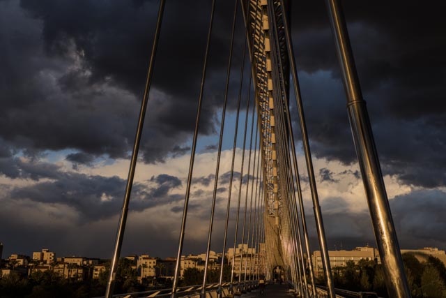 A bridge with sleek, modern cables stretches across a cityscape under a dramatic, stormy sky.