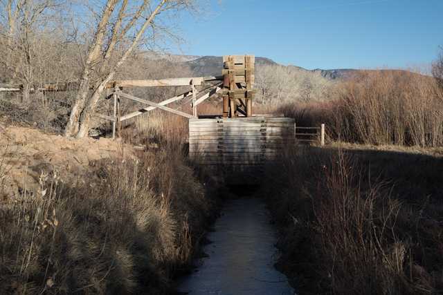 An old wooden waterwheel is situated atop a raised platform surrounded by tall grasses and trees.