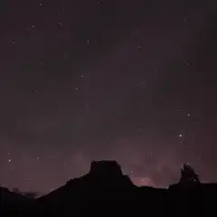 A night sky filled with stars, highlighted by the Milky Way galaxy, set against silhouetted mountain peaks.