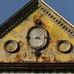 A weathered, triangular facade has a circular ornamental design centered, flanked by two circular recessed shapes and rust stains throughout a yellow surface.