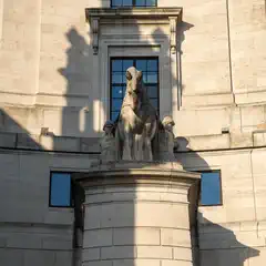 A stone sculpture of a horse flanked by two figures stands on a pedestal against a light-colored stone building with large windows.