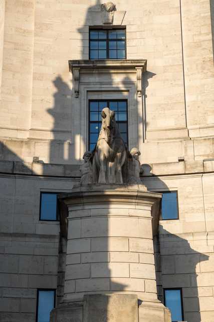 A stone sculpture of a horse flanked by two figures stands on a pedestal against a light-colored stone building with large windows.