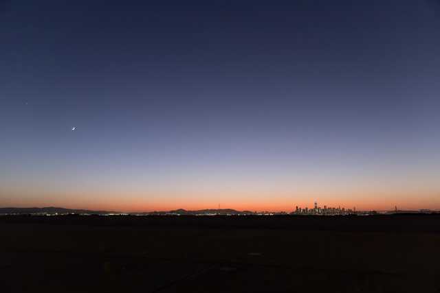A city skyline at dusk with a bridge and tower silhouetted against the fading light of day.