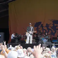 A crowd of people with raised hands and cameras, watching a man on stage playing a guitar, at an outdoor event.