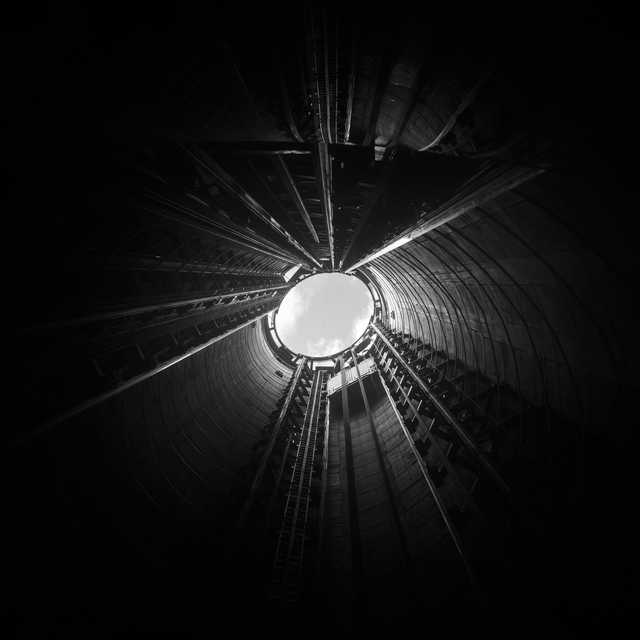 A circular opening at the top of a tall, dark industrial chimney reveals a bright sky, with radial metal structures and ladders converging toward the light.