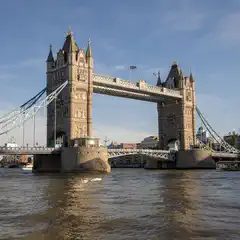 A stone bridge with two towers and a raised central span spans a wide river, with boats passing beneath and buildings visible along the far bank.