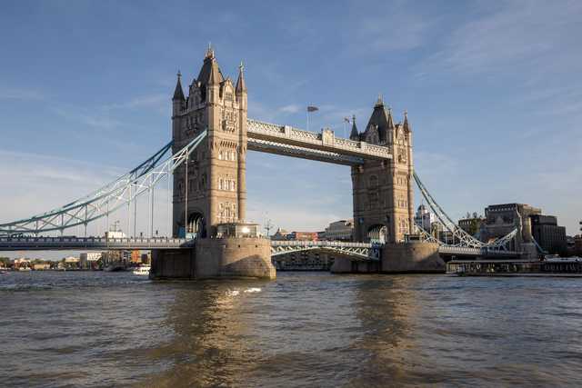 A stone bridge with two towers and a raised central span spans a wide river, with boats passing beneath and buildings visible along the far bank.