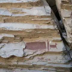 A large rock formation features a red pictograph depicting a human figure standing next to a deer, surrounded by additional handprints and symbols.