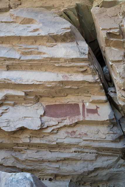 A large rock formation features a red pictograph depicting a human figure standing next to a deer, surrounded by additional handprints and symbols.