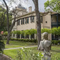 A large, cream-colored house surrounded by trees and a statue in the foreground.