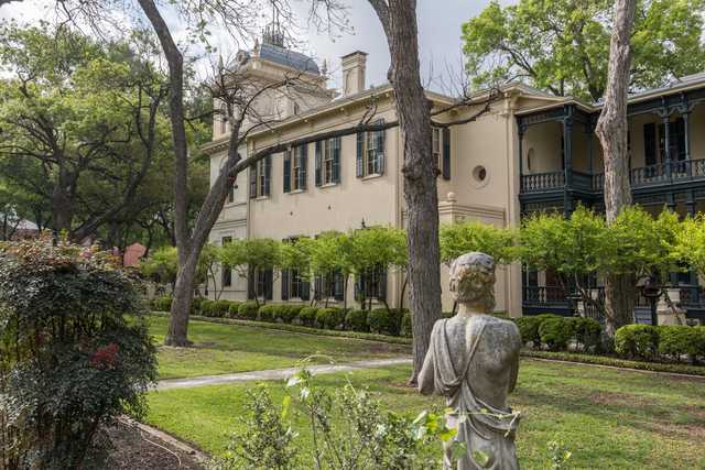 A large, cream-colored house surrounded by trees and a statue in the foreground.