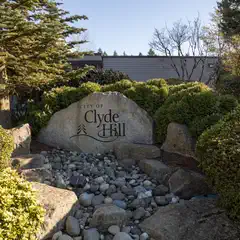 A sign sits behind a collection of rocks, surrounded by trees and bushes on either side. "City of Clyde Hill" is engraved in black text on the front of the stone.