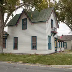 A white house with blue trim and green roof is situated on a grassy lawn surrounded by trees.