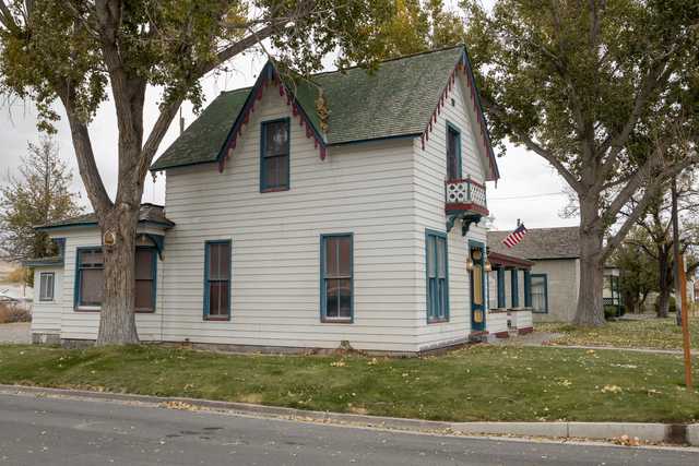 A white house with blue trim and green roof is situated on a grassy lawn surrounded by trees.