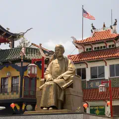 A golden statue of a man sitting on a throne in front of an oriental building with tiled roof and red lanterns hanging from poles.