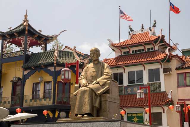 A golden statue of a man sitting on a throne in front of an oriental building with tiled roof and red lanterns hanging from poles.
