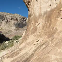 The moon hangs low in a blue sky over a rocky cliff face with a desert landscape below.