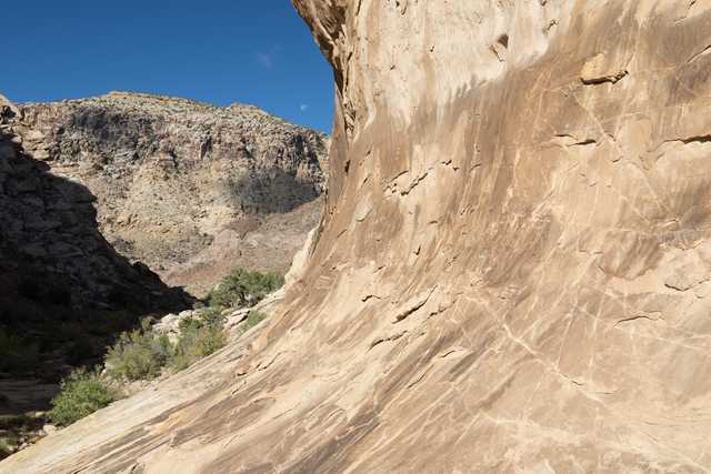 The moon hangs low in a blue sky over a rocky cliff face with a desert landscape below.