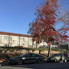 A white building with a brown roof and arched windows sits atop a concrete retaining wall, behind which is a grassy area enclosed by a chain link fence.