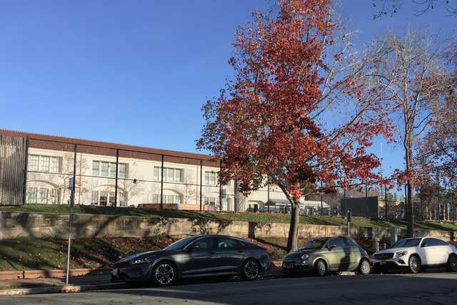 A white building with a brown roof and arched windows sits atop a concrete retaining wall, behind which is a grassy area enclosed by a chain link fence.