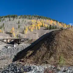 A pile of dirt and rocks next to a bridge in front of mountains with trees that are changing color for fall.