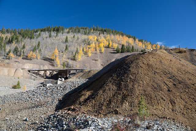 A pile of dirt and rocks next to a bridge in front of mountains with trees that are changing color for fall.