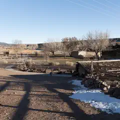 A dirt path leads past a pile of rusted farm equipment and a wooden fence to a small log cabin with a sloping roof.