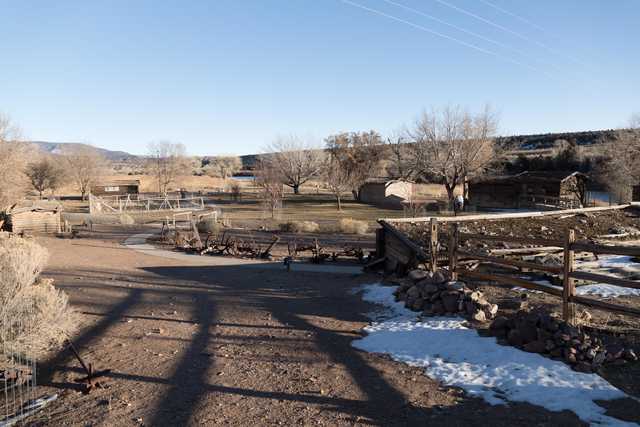 A dirt path leads past a pile of rusted farm equipment and a wooden fence to a small log cabin with a sloping roof.