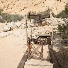 A wooden structure with a metal cage rests on a sandy surface before a rock hillside with sparse vegetation.