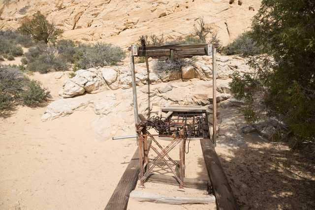 A wooden structure with a metal cage rests on a sandy surface before a rock hillside with sparse vegetation.