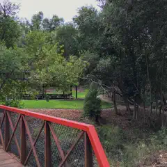 A red metal bridge with a wooden deck crossing over a creek surrounded by trees and grass.