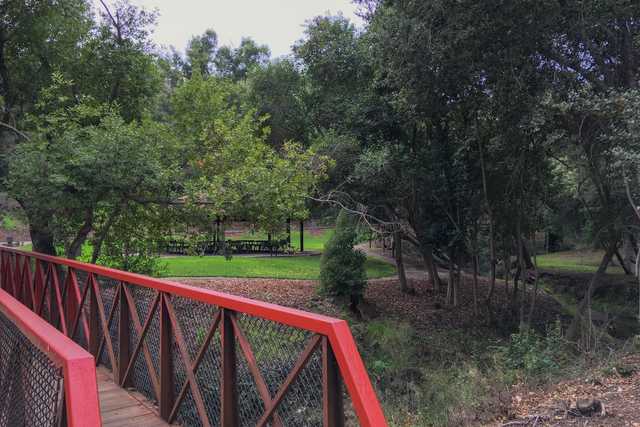 A red metal bridge with a wooden deck crossing over a creek surrounded by trees and grass.