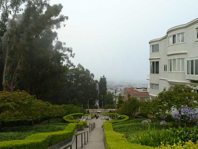 A white building stands adjacent to a terraced garden with a staircase leading down toward rooftops and a distant, hazy landscape.