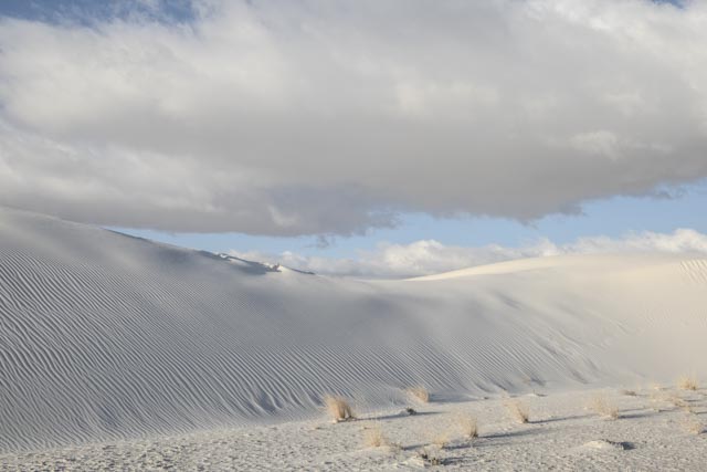 An expanse of white sand dunes stretches across the landscape under a blue sky with clouds.