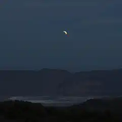 A crescent moon hangs above mountains and a body of water at night.
