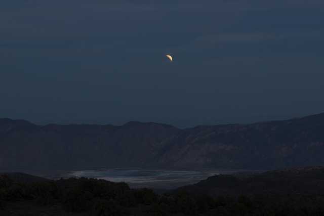 A crescent moon hangs above mountains and a body of water at night.