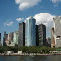 A row of skyscrapers stands behind a waterfront promenade with trees and a body of water in the foreground.