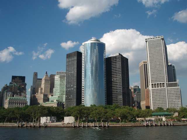 A row of skyscrapers stands behind a waterfront promenade with trees and a body of water in the foreground.