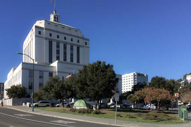 A large white building stands behind a grassy area with several trees and tents.