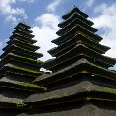 A thatched roofed building with several tiers, topped with gold ornamentation, sits beneath a blue sky filled with white clouds.