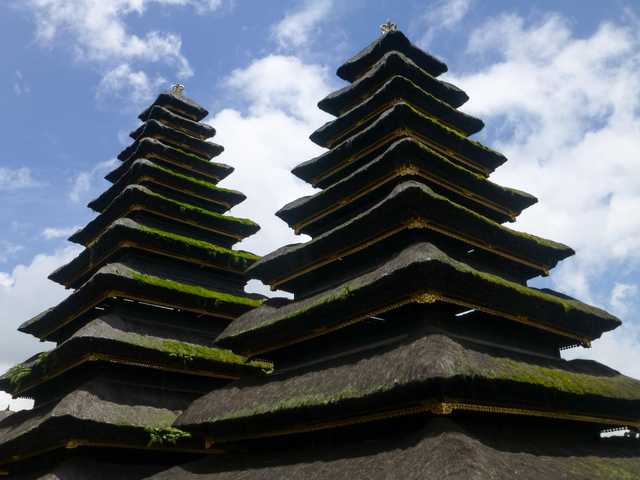 A thatched roofed building with several tiers, topped with gold ornamentation, sits beneath a blue sky filled with white clouds.