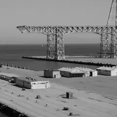 A large industrial crane stands over a dock with several small buildings and a fence, overlooking calm water under an overcast sky.