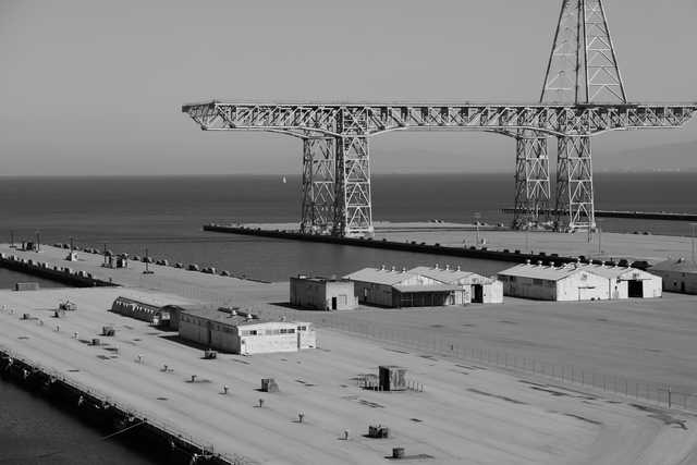 A large industrial crane stands over a dock with several small buildings and a fence, overlooking calm water under an overcast sky.