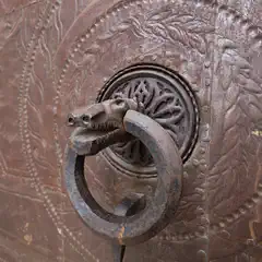 A metal door handle in the shape of a horse's head is prominently featured on a rusty metal door with ornate leaf designs.