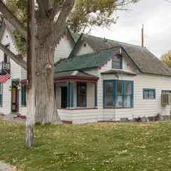 A white house with blue trim and a green roof, featuring an American flag on the porch, surrounded by trees and grass.