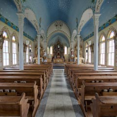 A church interior with high ceiling decorated in blue and gold, wooden pews, and stained glass windows providing natural light.