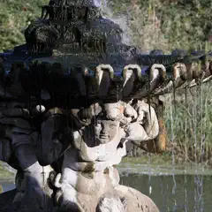 Water cascades from a moss-covered stone fountain supported by sculpted figures, with greenery and a still pool in the background.