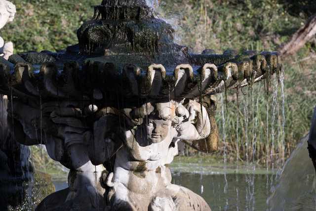Water cascades from a moss-covered stone fountain supported by sculpted figures, with greenery and a still pool in the background.